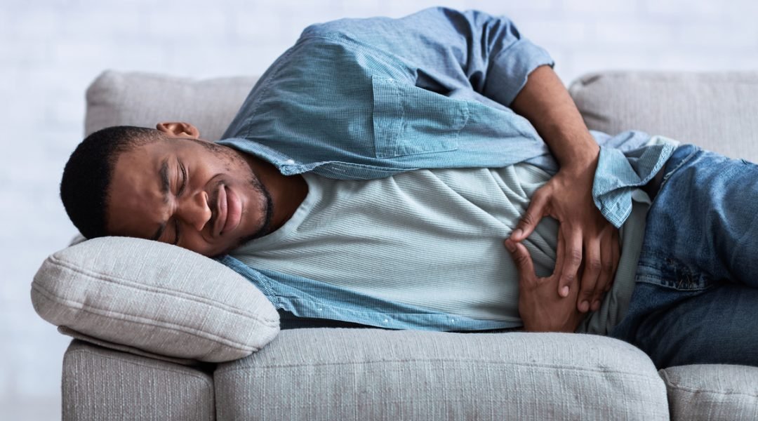 Stomachache. African Guy Suffering From Abdominal Pain Touching Aching Stomach Lying On Sofa At Home. Black Man Having Gastritis Problem And Abdomen Inflammation. Health Issue Concept